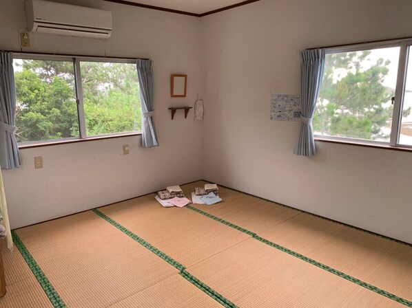 Ein Zimmer mit Tatami-Boden, zwei Fenstern mit Vorhängen, einer Wand-Klimaanlage und einem kleinen Regal mit einem gerahmten Bild.