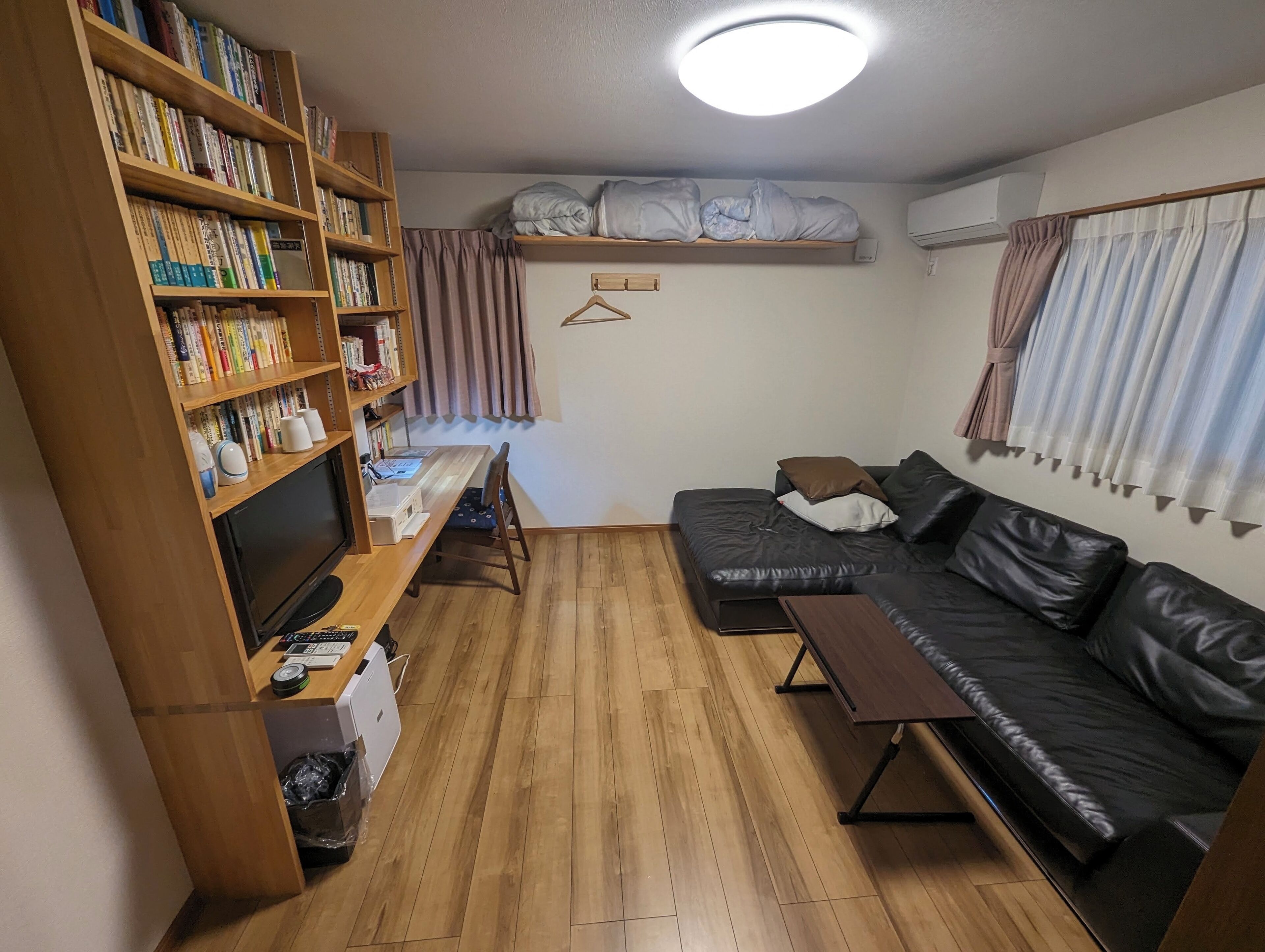 A cozy living room with a wooden bookshelf, a black leather sofa, a wooden coffee table, and a TV.
