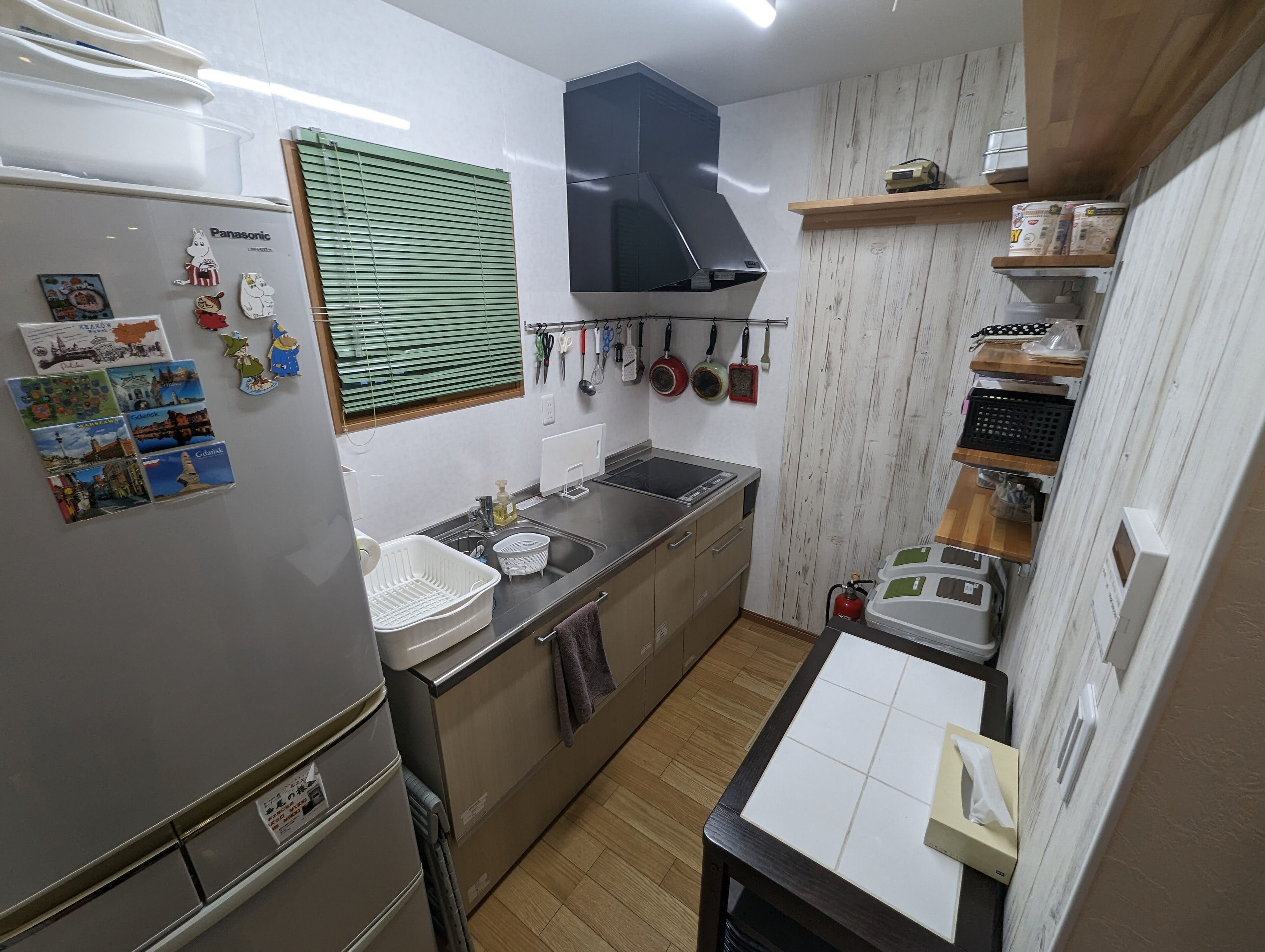 A compact kitchen with a refrigerator, sink, and stove. There are various kitchen utensils hanging on the wall, a window with blinds, and a shelf with kitchen items.