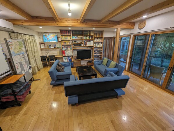 A spacious living room with wooden beams, a large bookshelf, a flat-screen TV, and a large window with a view of the outdoors. - guesthouse KEY's (Nishio)