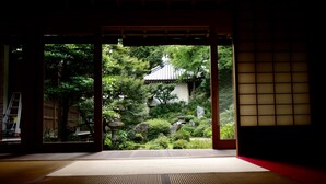 Una habitación tradicional japonesa con suelo de tatami, una puerta corredera shoji y vistas a un jardín con un edificio y árboles.