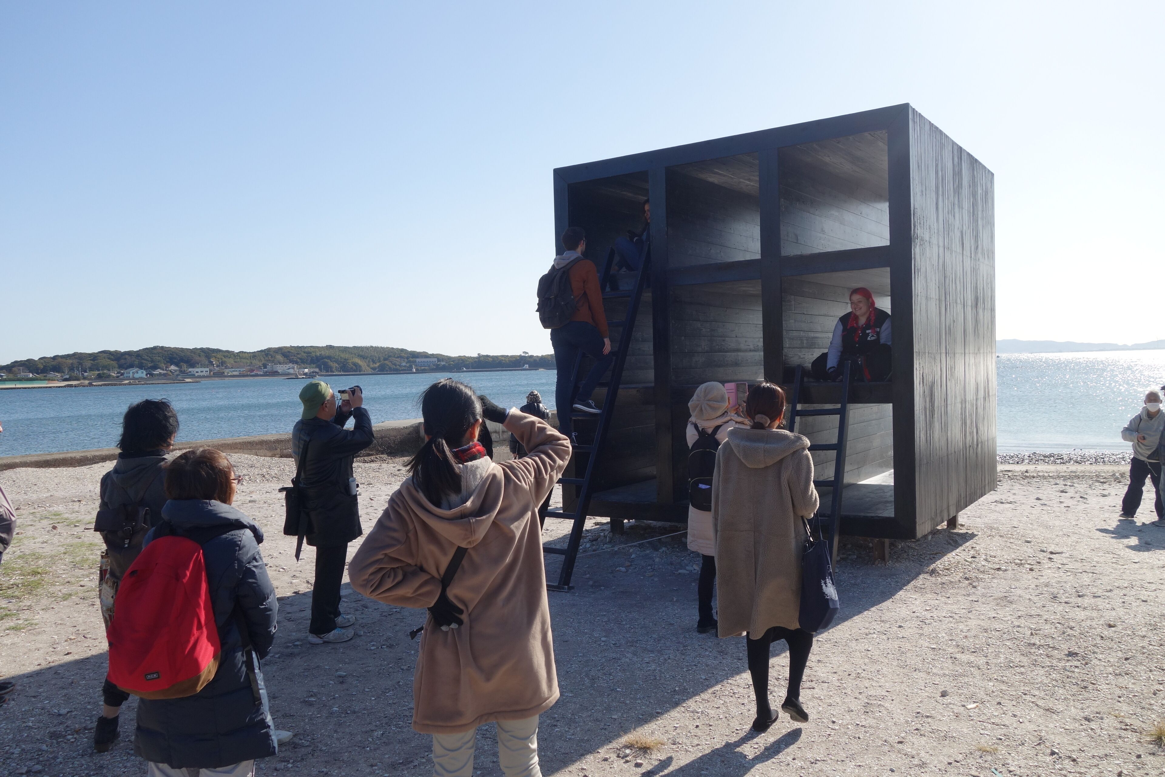 A modern, minimalist building with a glass facade, situated on a beach with a clear view of the sea and distant hills.