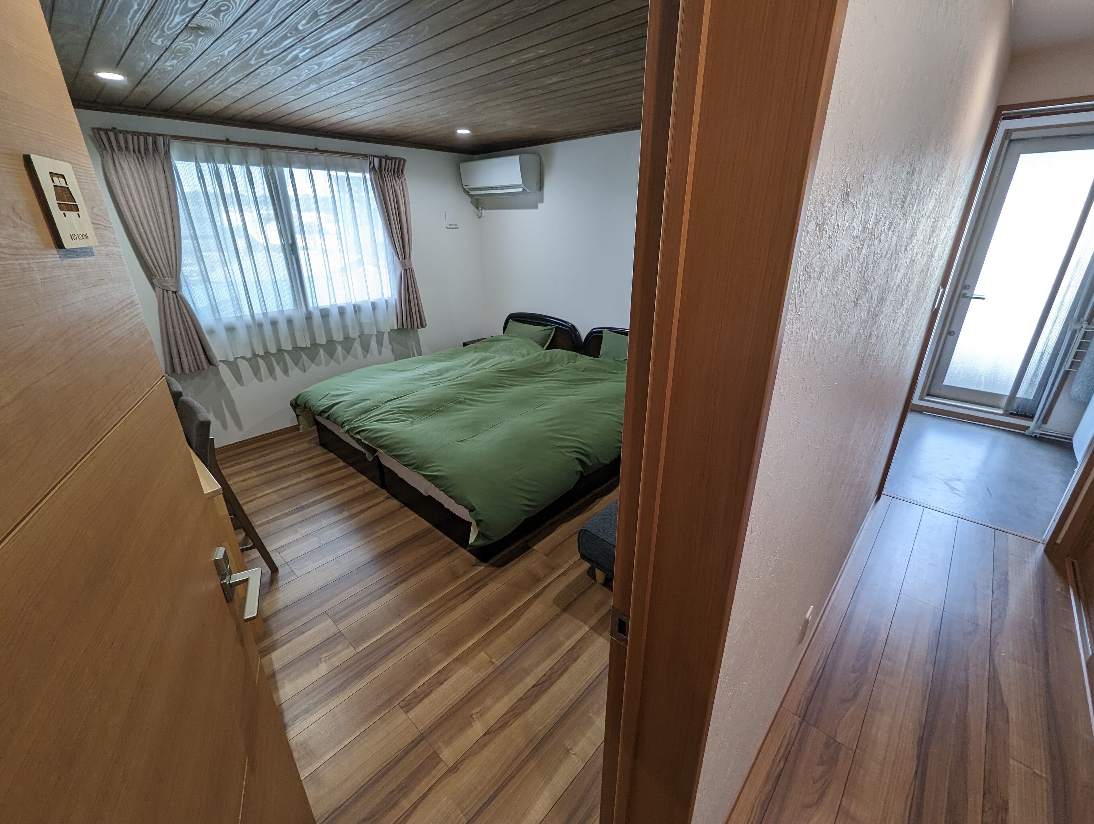 Ein Schlafzimmer mit Holzboden, einem Bett mit grüner Bettwäsche, einem Fenster mit Vorhängen und einer Klimaanlage an der Wand.