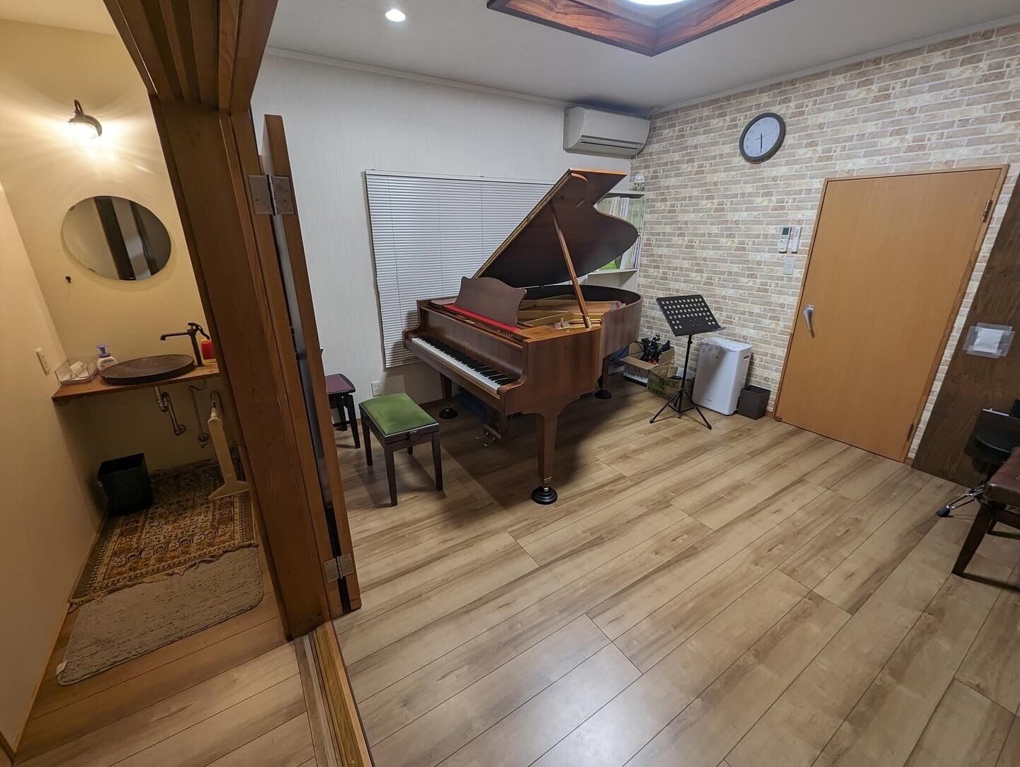 A room with a grand piano, a small stool, a mirror, a sink, and a clock on the wall.