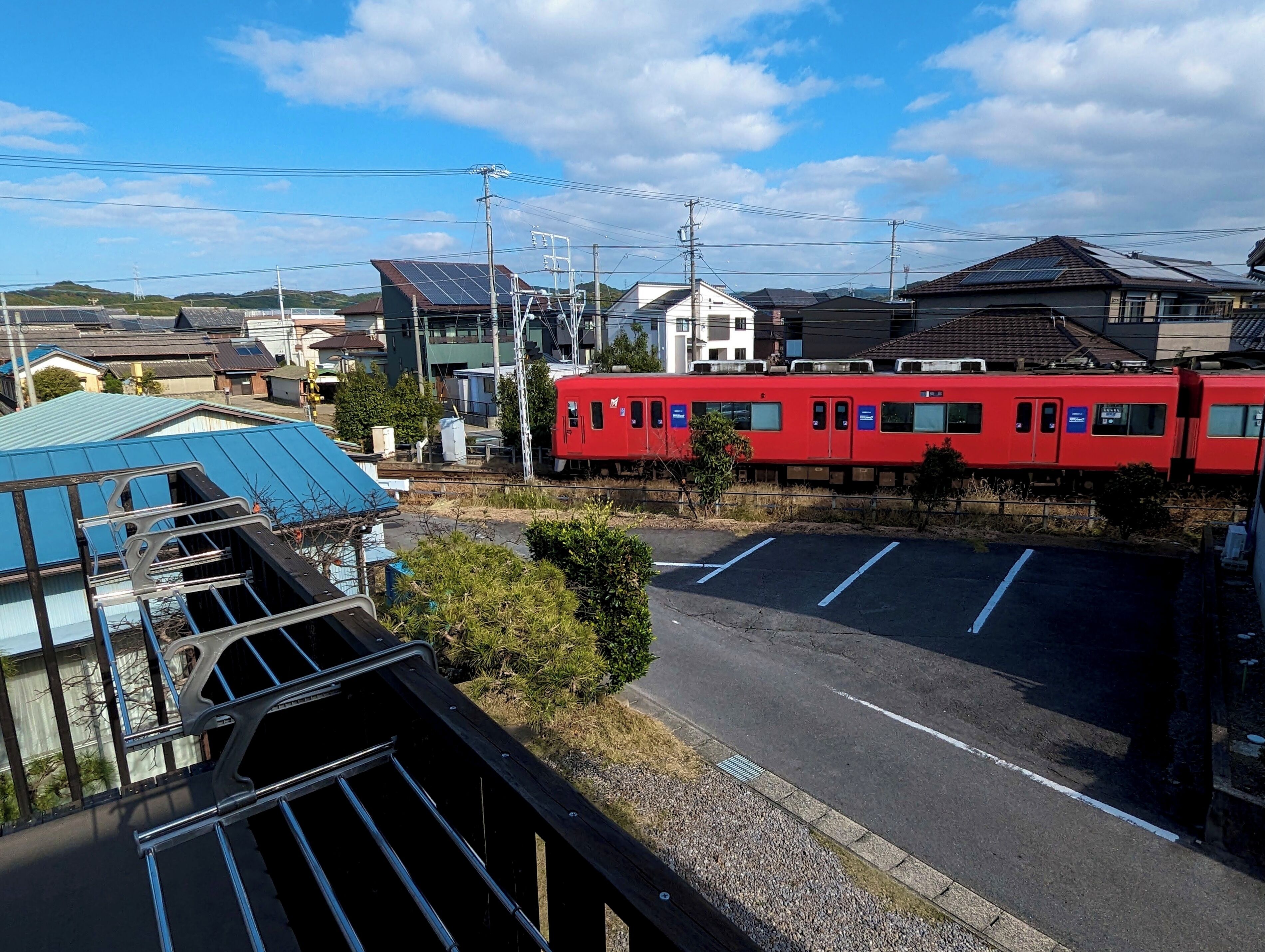 Un train rouge sur des rails, avec un parking et des bâtiments en arrière-plan.