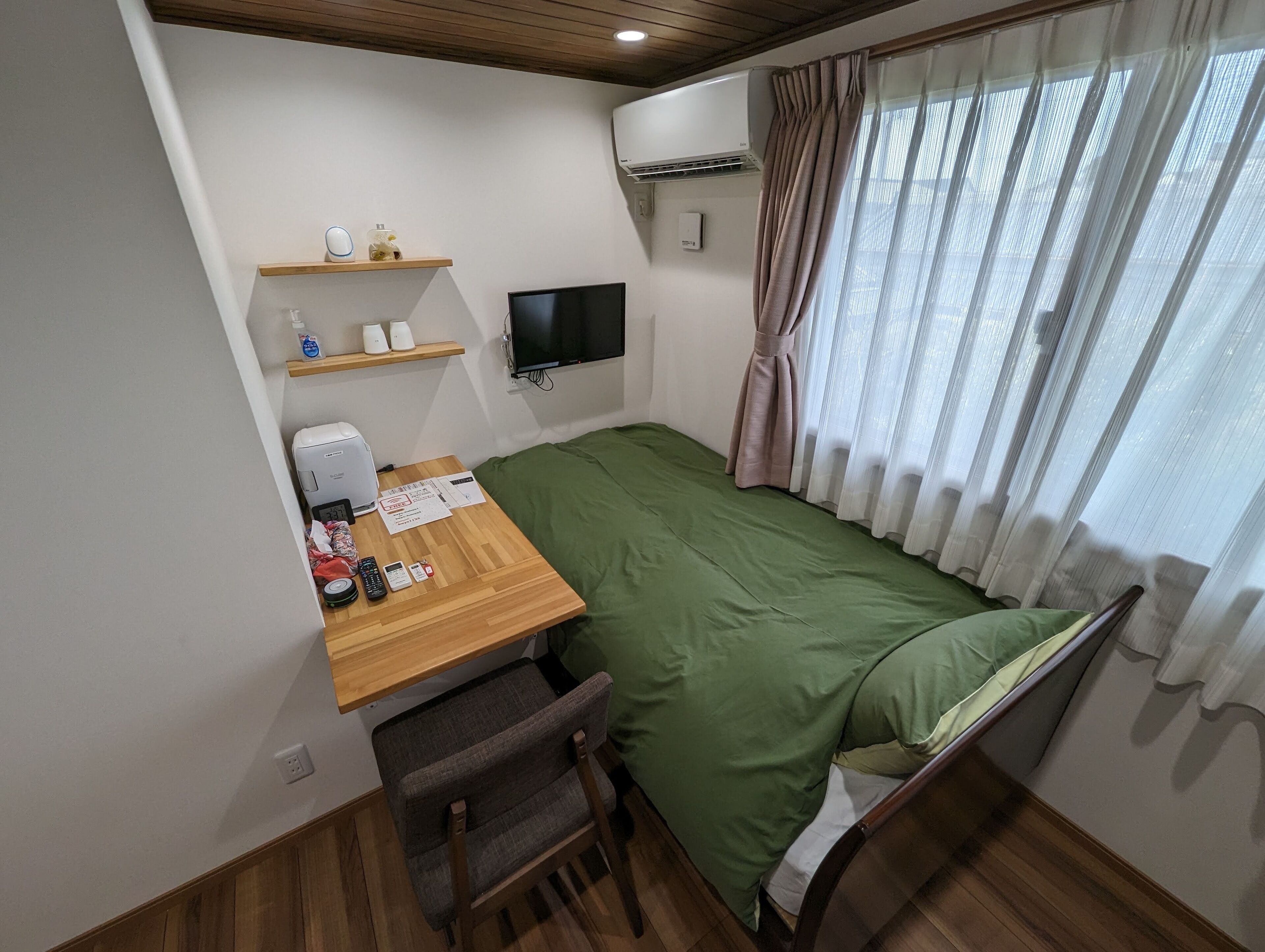 A compact room with a bed, a desk, a chair, a TV, and a window with curtains.