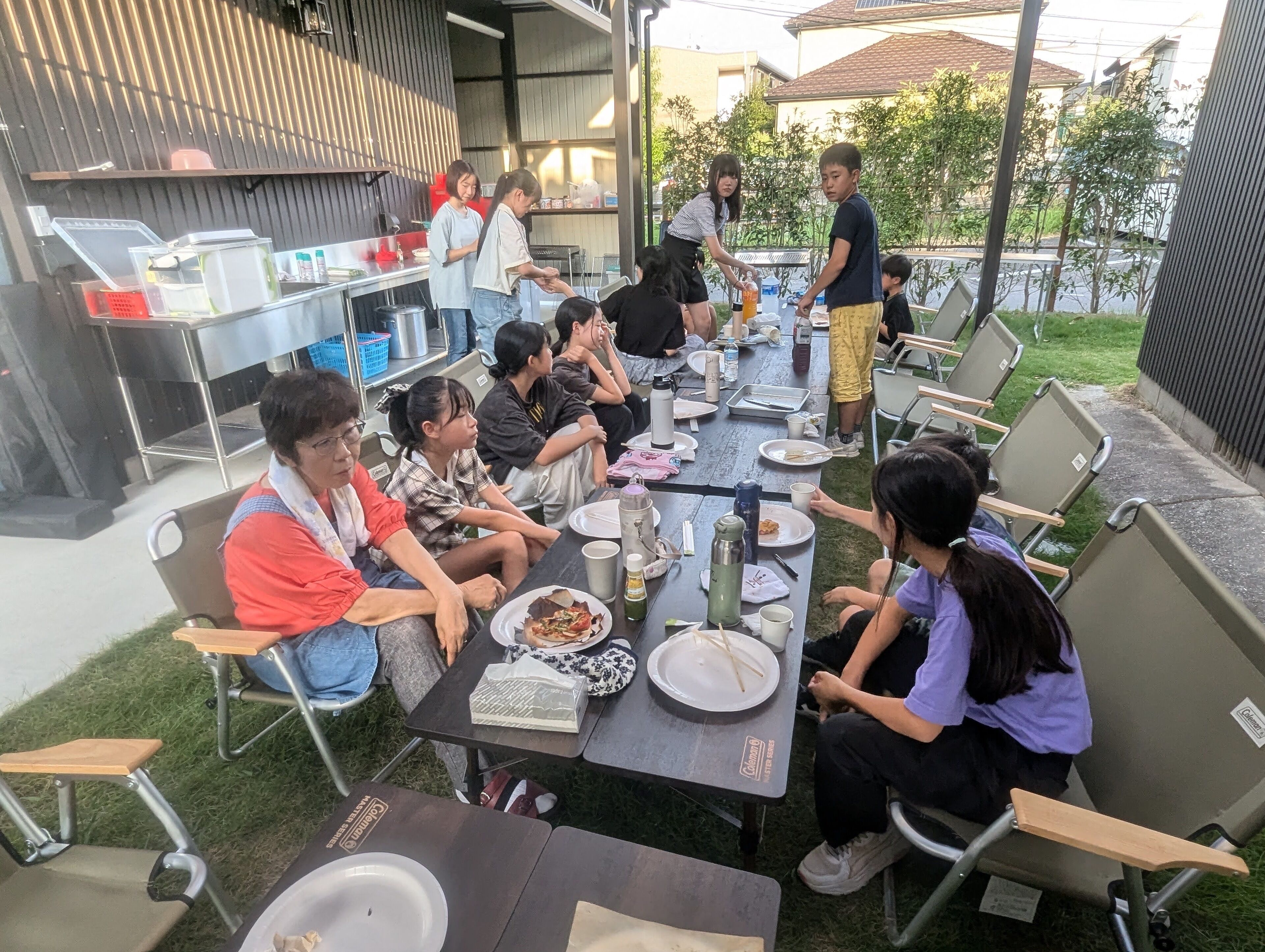 Un'area esterna con tavoli e sedie, una postazione barbecue e un gruppo di persone che mangiano.