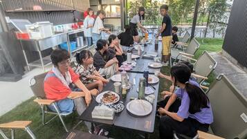 Un'area esterna con tavoli e sedie, una postazione barbecue e un gruppo di persone che mangiano.