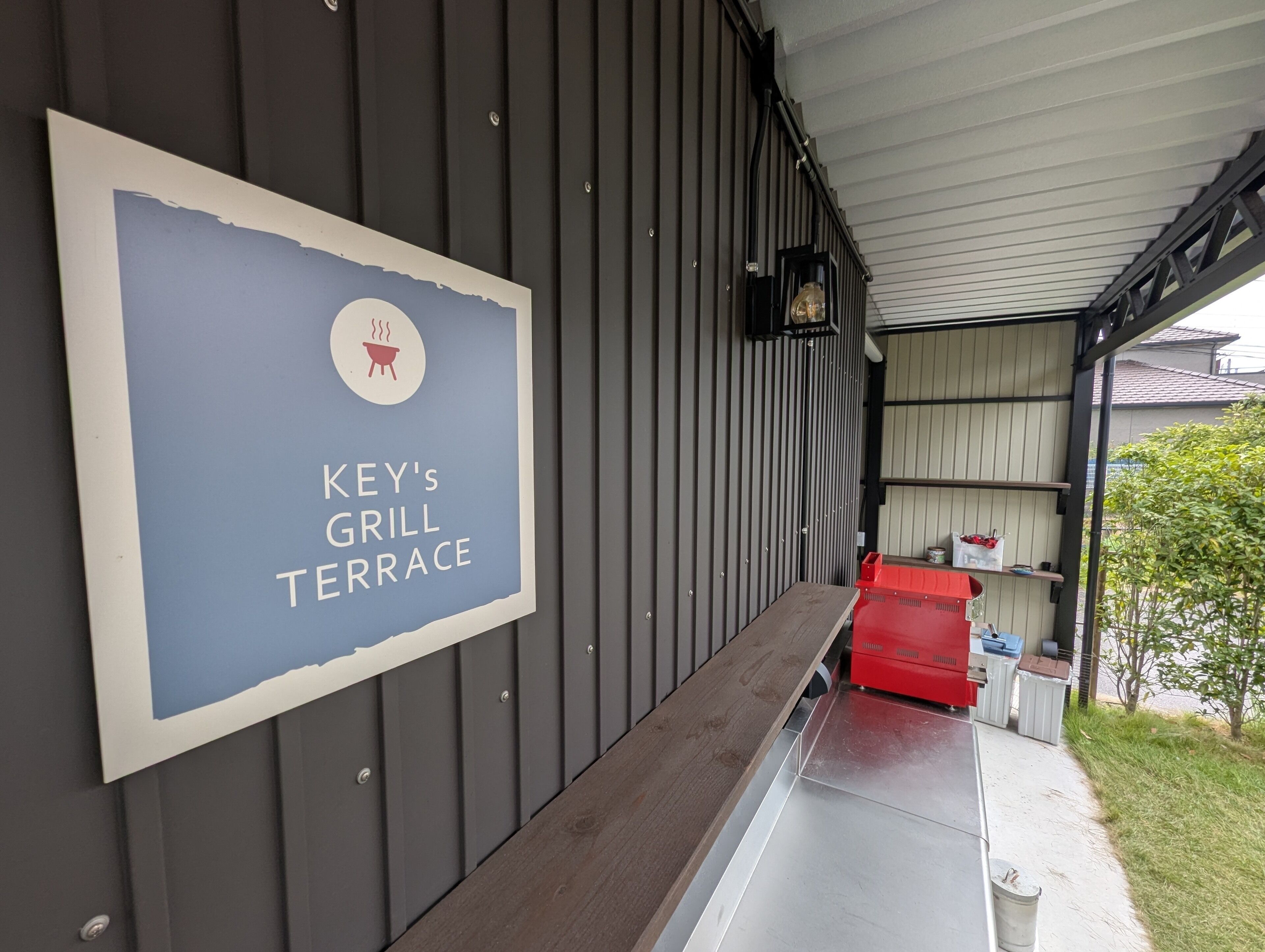 A sign for "Key's Grill Terrace" with a barbecue grill icon, a wooden bench, and a red trash bin outside a building with a metal exterior.