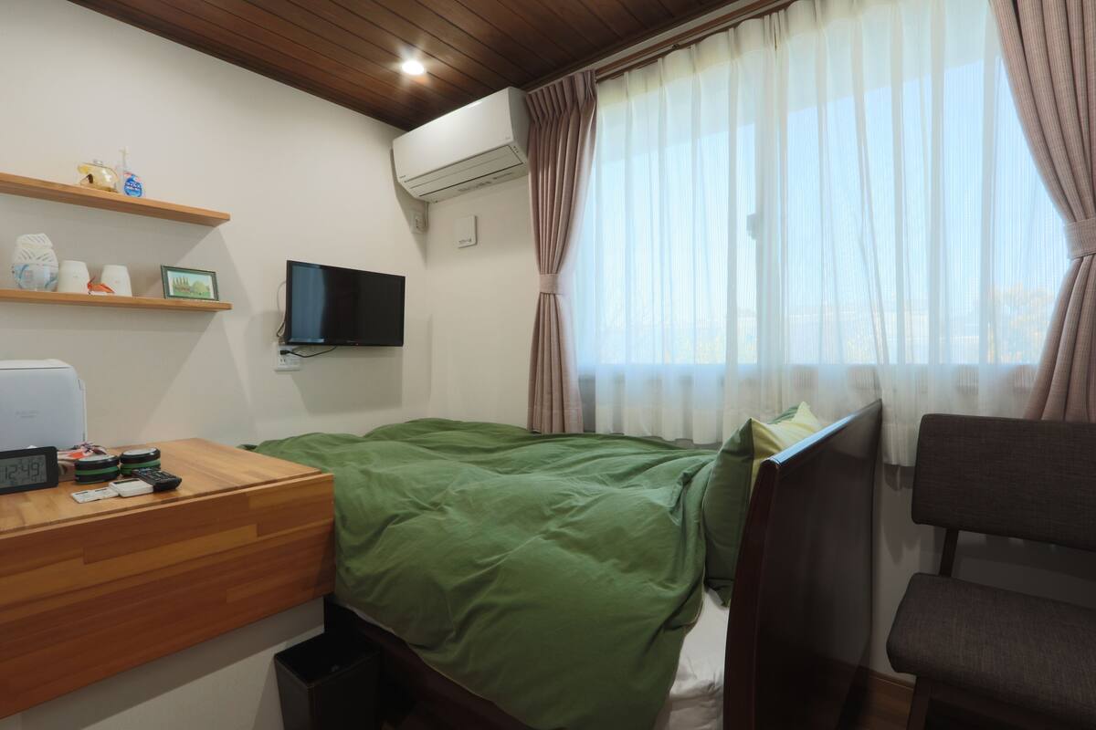 A bedroom with a bed, a desk with a clock, a TV, and a window with curtains.