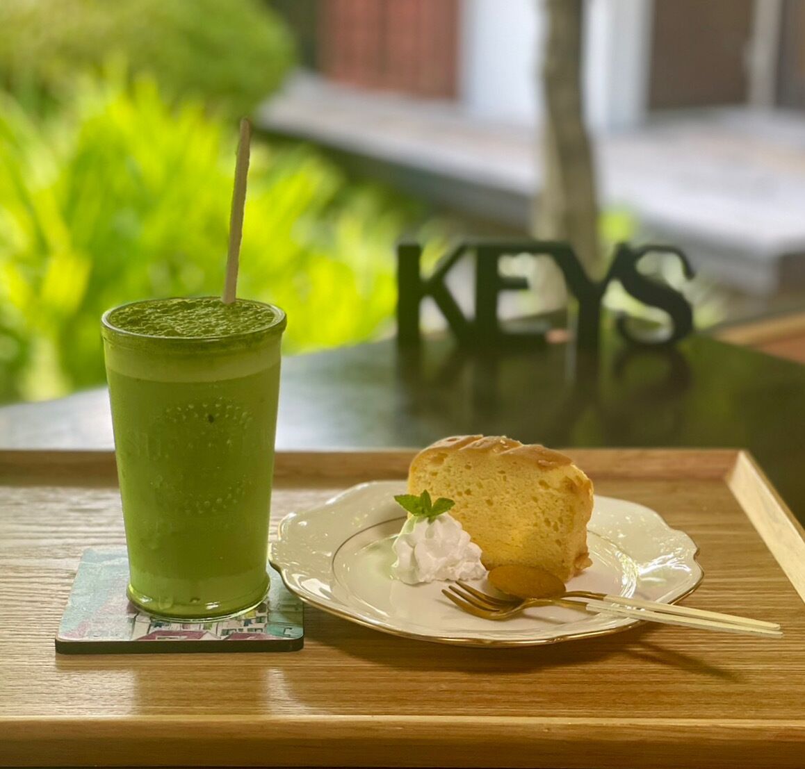 Una bebida verde en un vaso con pajita, una rebanada de pastel en un plato con un tenedor, y una mesa de madera con un posavasos.