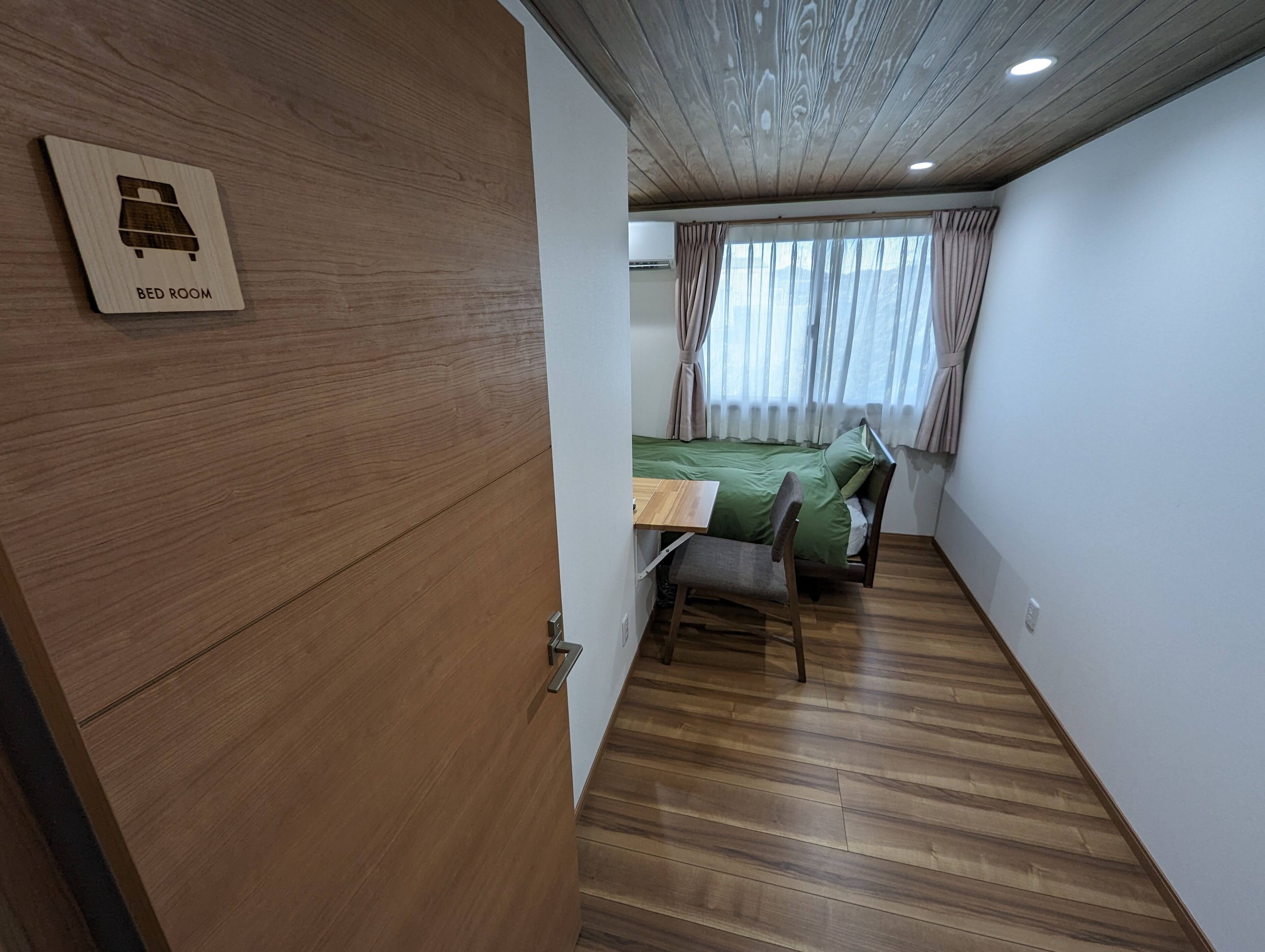 A compact room with a bed, a desk, and a chair. A wooden door with a bed icon is visible.