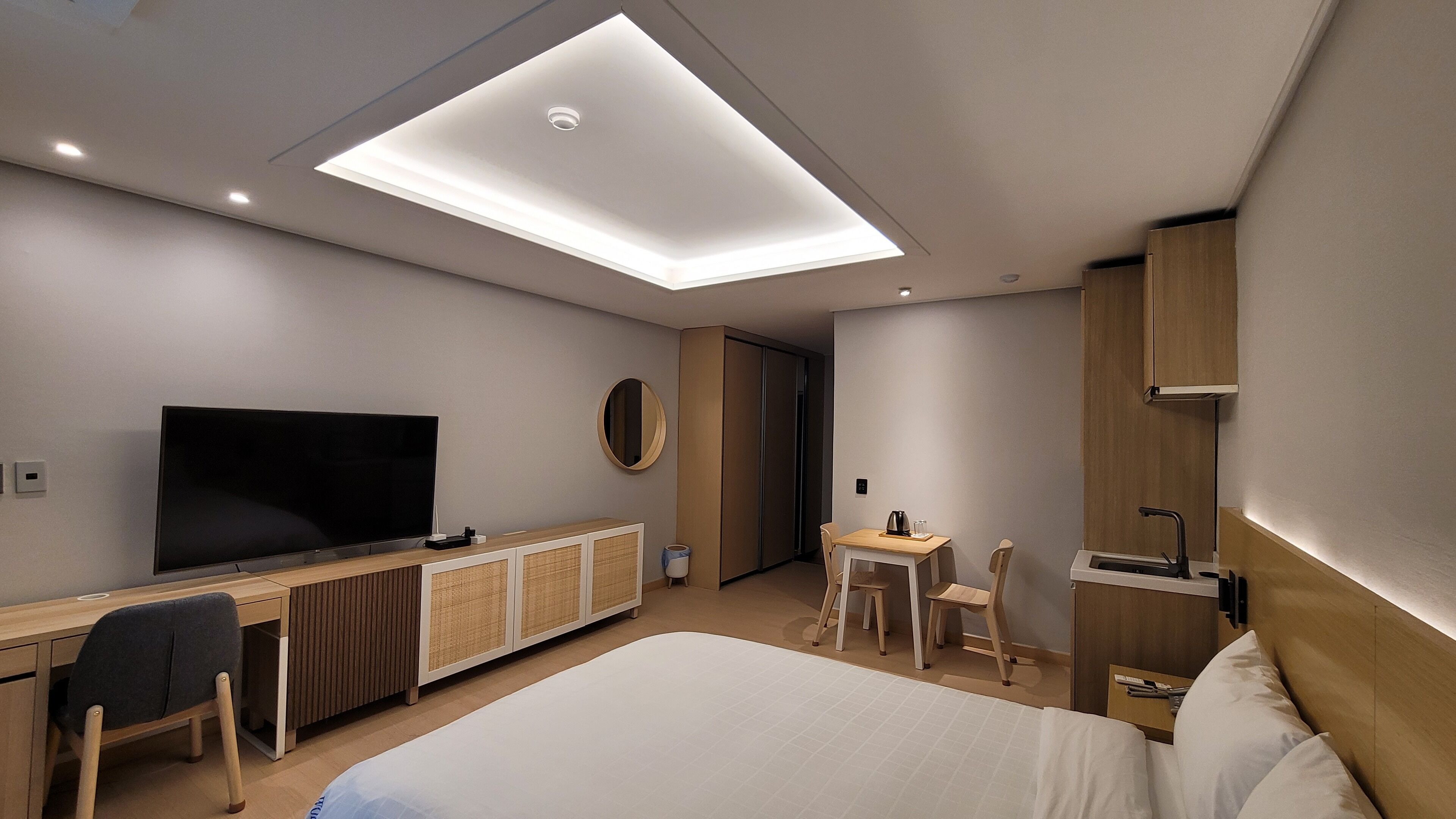 A modern hotel room with a large bed, a desk with a chair, a television, a small table with a chair, and a wardrobe.