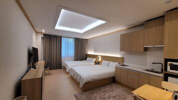 A hotel room with a bed, a desk, a TV, a ceiling light, a window with curtains, and a kitchenette.