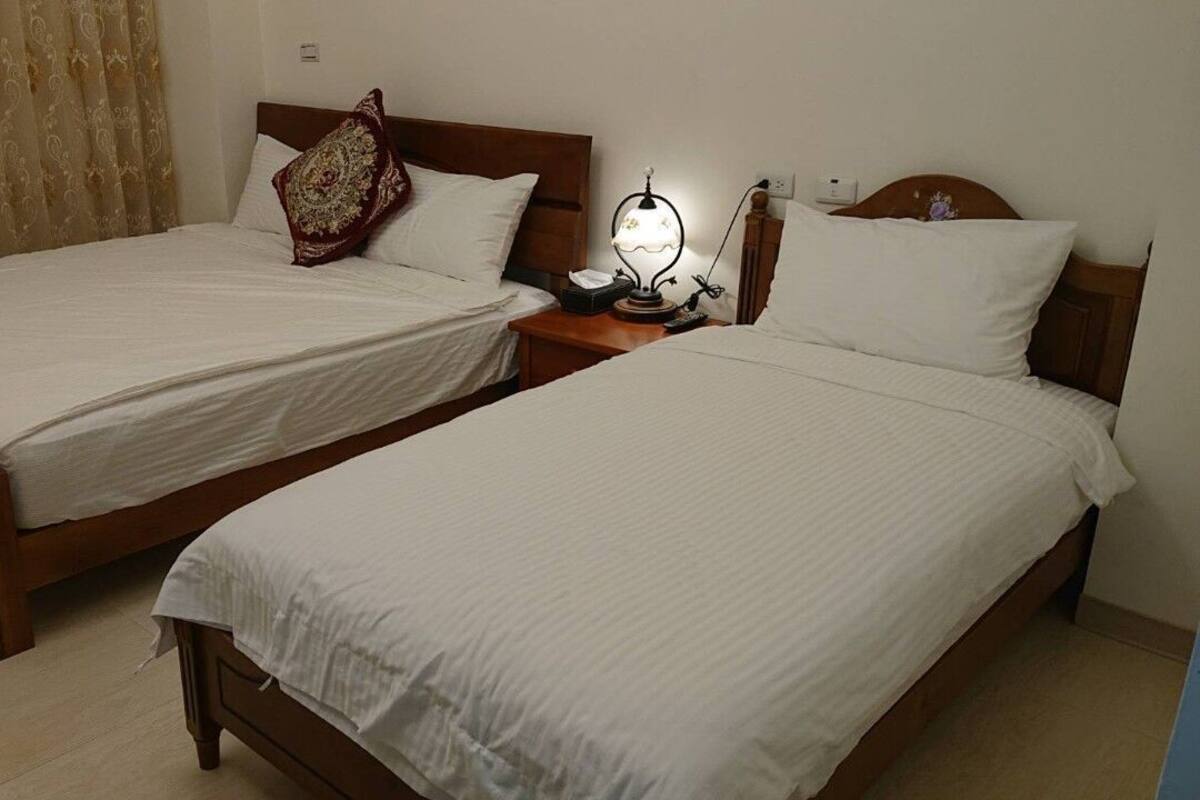 Two single beds with wooden frames and white linens, a bedside table with a lamp, and a patterned curtain.