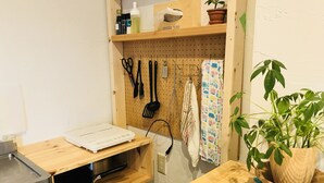 Une cuisine avec des étagères en bois, une plaque de cuisson à induction blanche, une casserole, une poêle, un porte-ustensiles suspendu, une plante en pot et un set de table bleu.