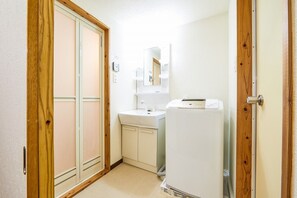 A small, compact bathroom with a wooden door, a white sink, a mirror, and a white washing machine. - Yanbaru Forest Hills (Motobu)