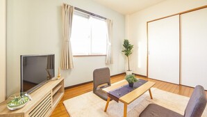 A modern living room with a flat-screen TV, two chairs, a small table, a potted plant, and a sliding door. - Yanbaru Forest Hills (Motobu)