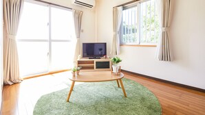 A room with a wooden floor, a green rug, a TV on a stand, and two windows with curtains. - Yanbaru Forest Hills (Motobu)
