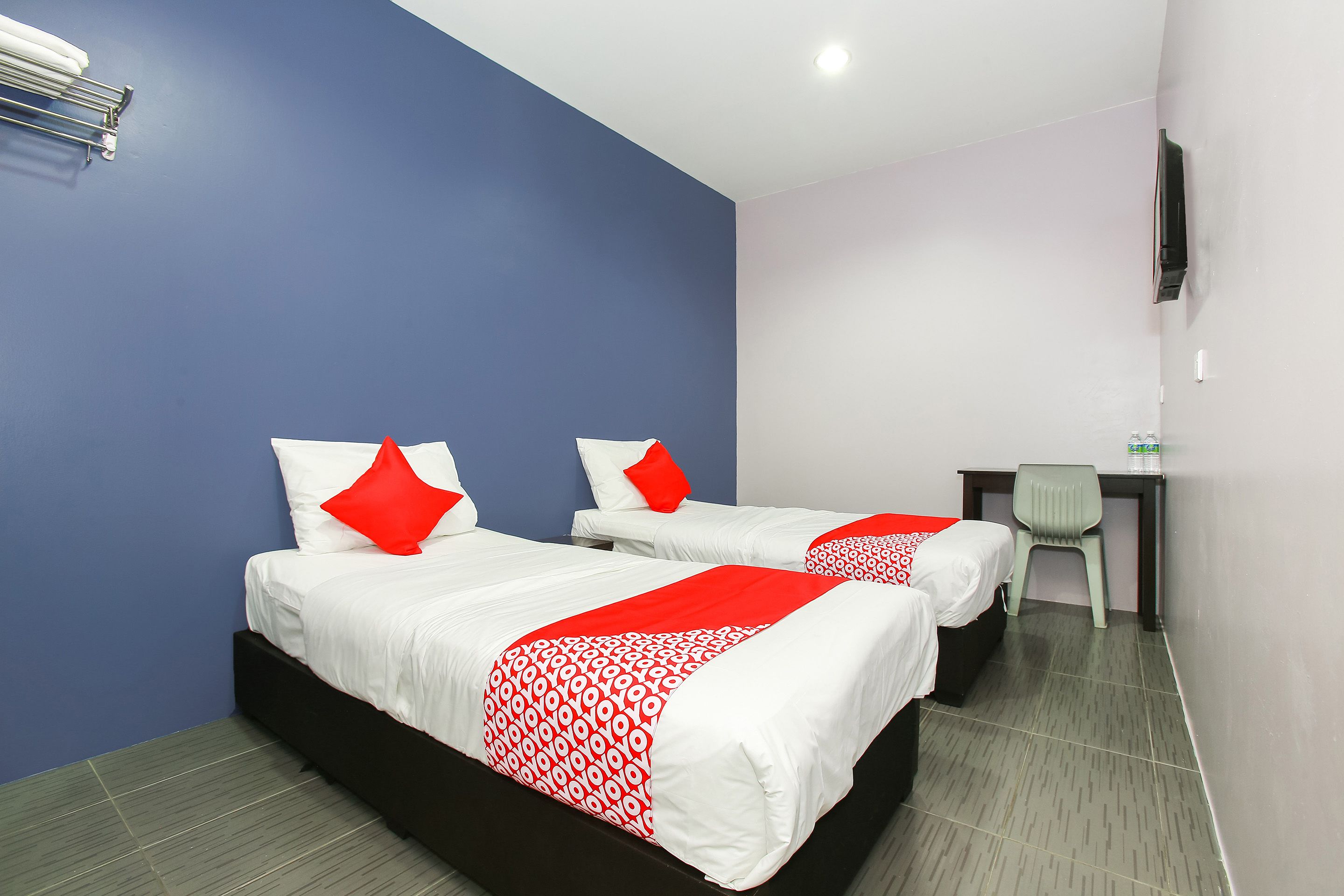 A hotel room with two beds, each with white bedding and red accents, a desk with a chair, and a flat-screen TV mounted on the wall.