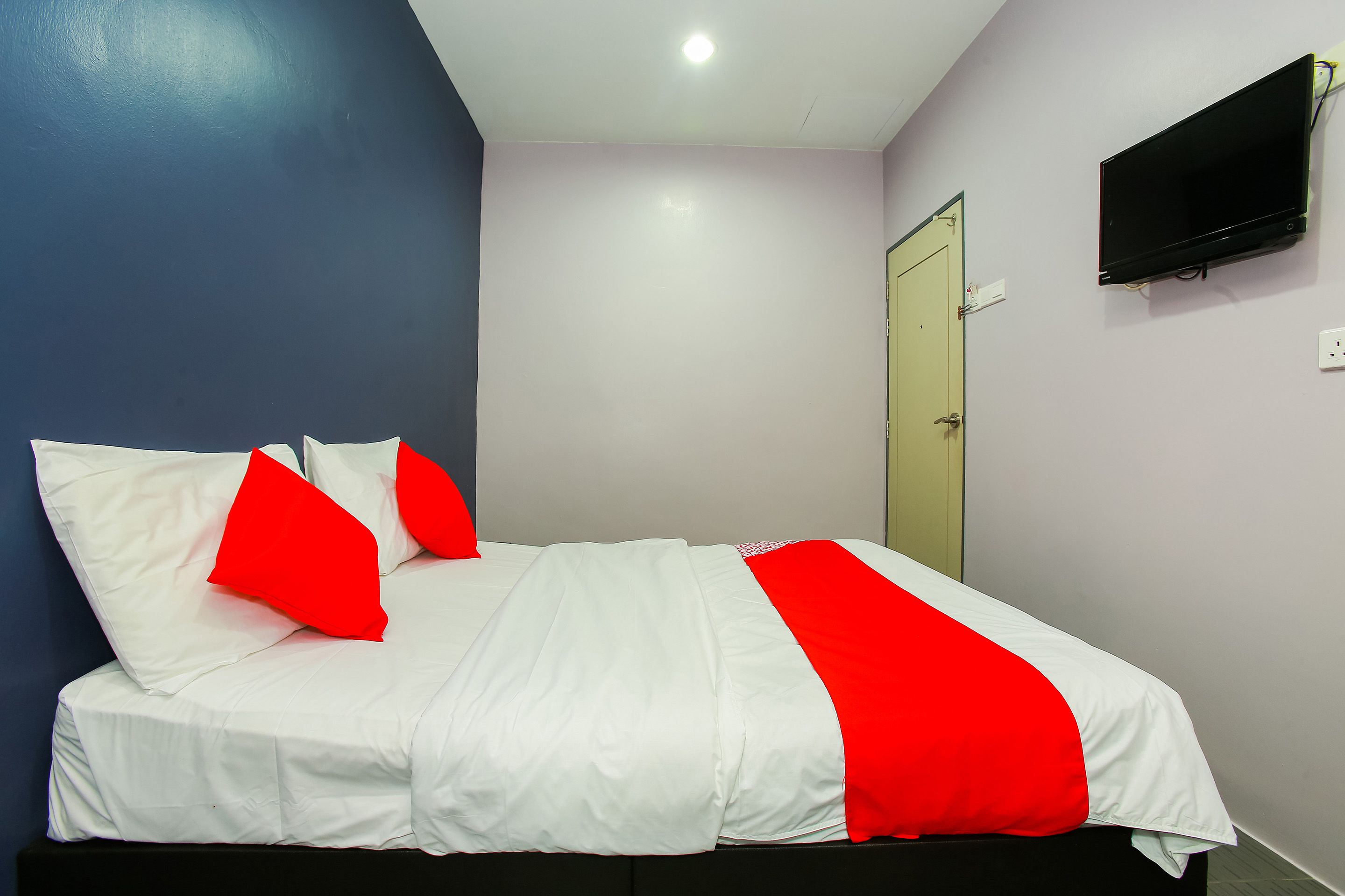 A bed with white and red bedding, a flat-screen TV mounted on the wall, and a door with a light switch.
