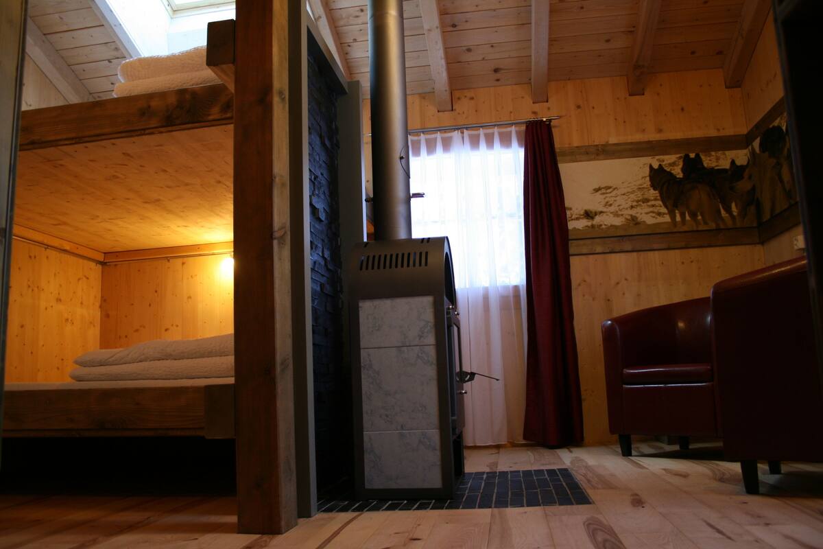 A wooden cabin interior with a bed, a fireplace, a red chair, and a wooden ceiling.