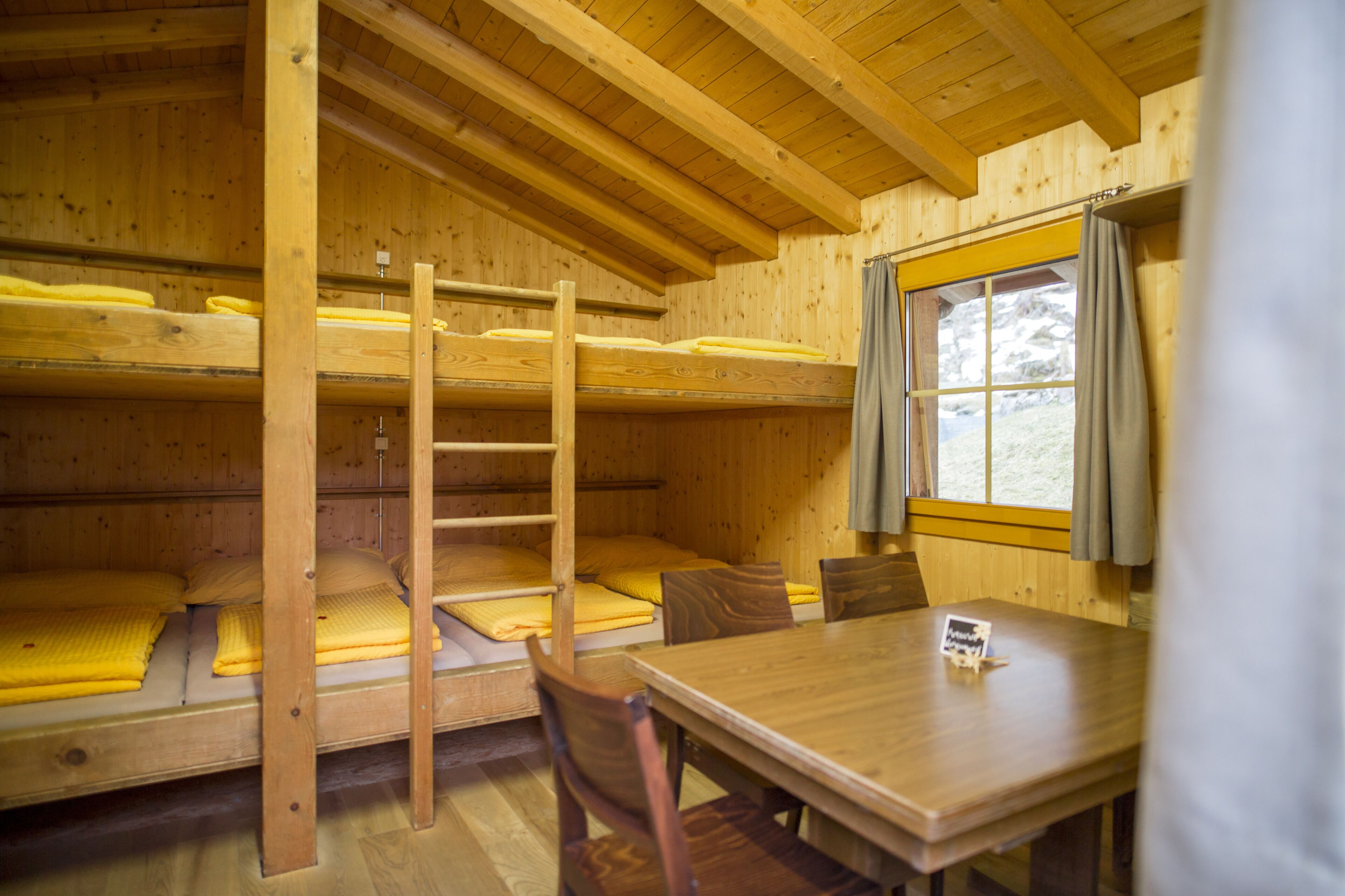 A wooden cabin interior with bunk beds, a dining table, and a window with curtains.