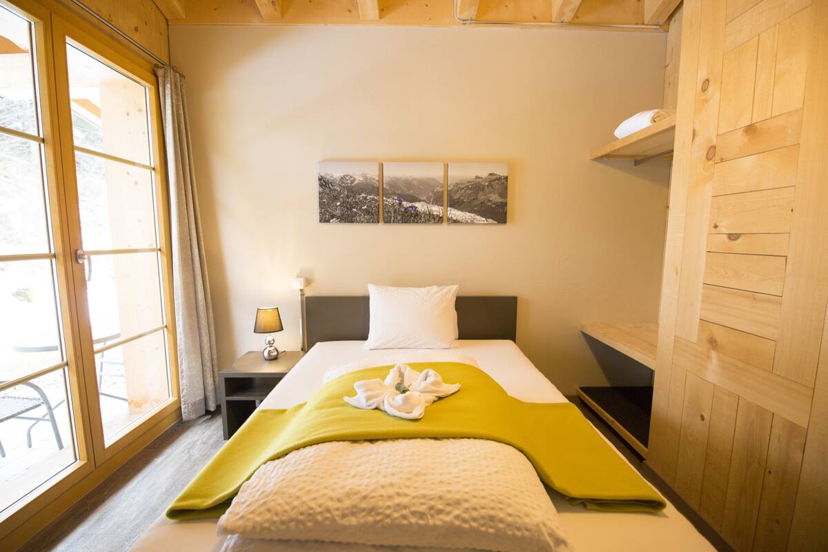 A hotel room with a bed, a nightstand, a lamp, a window with curtains, and a wall-mounted picture.
