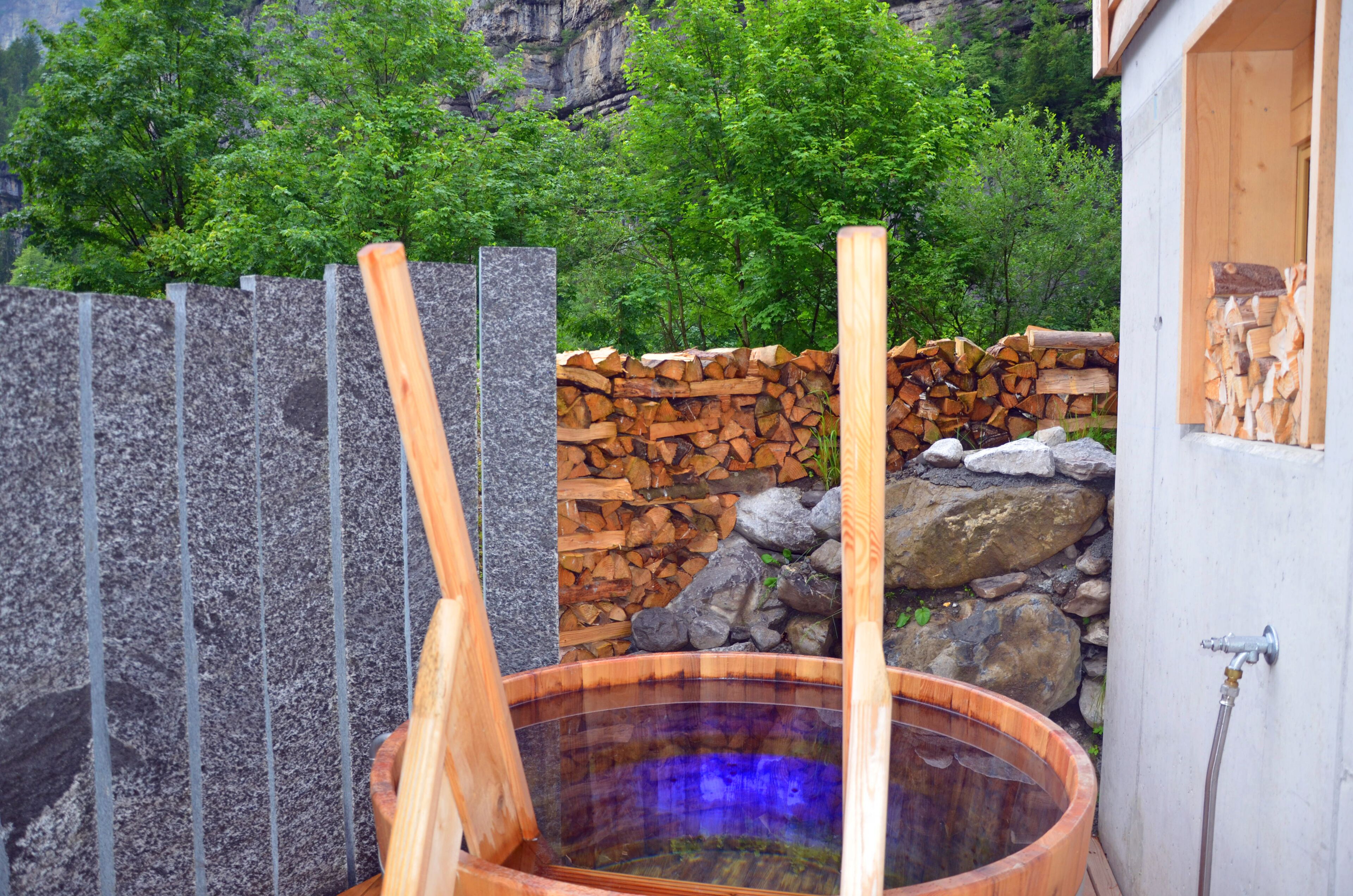 A hot tub with wooden benches, a stone wall, and a stack of firewood.