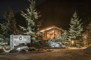 Un chalet au revêtement extérieur en bois et aux murs en verre, entouré d’arbres et de rochers couverts de neige.