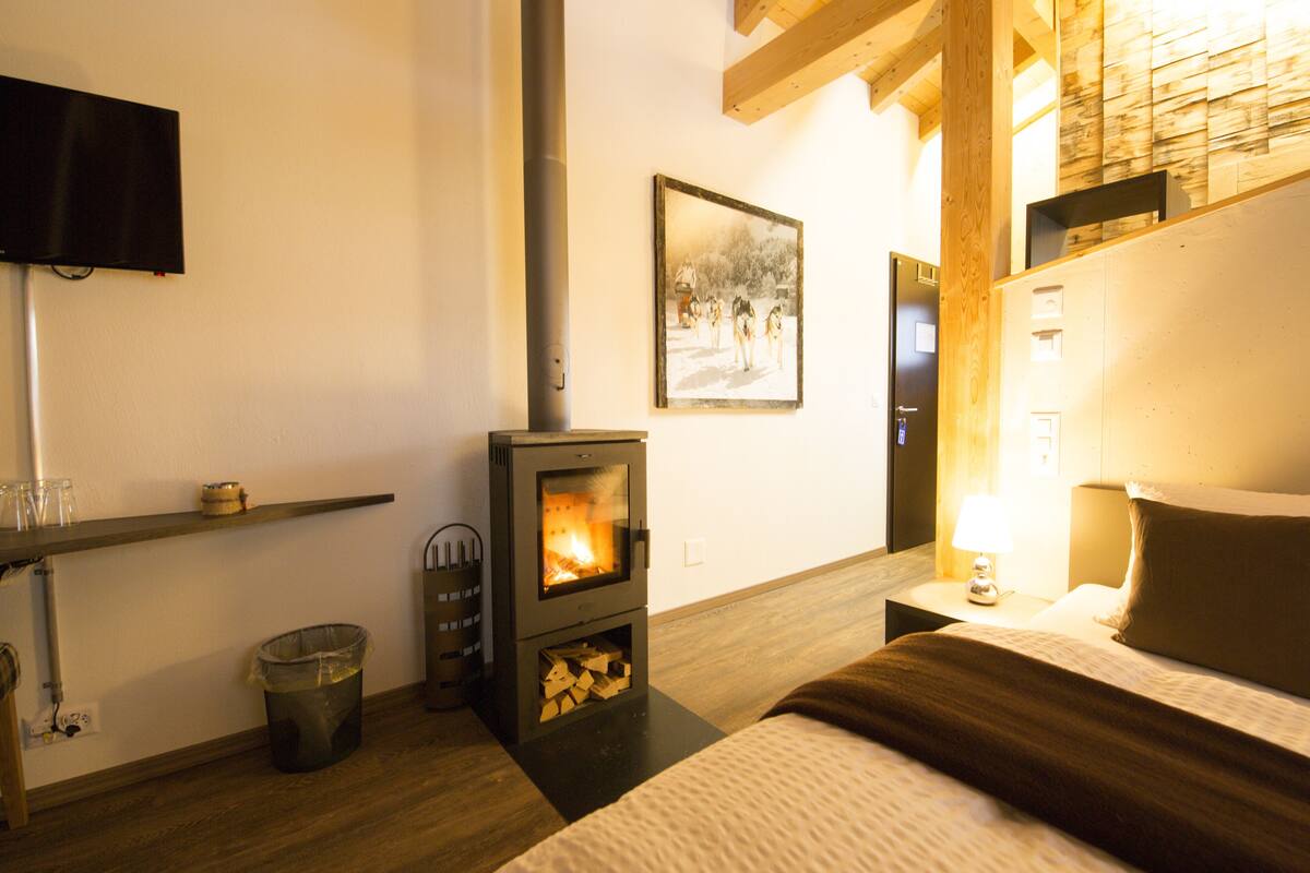 A cozy hotel room with a wood-burning stove, a bed, a bedside table with a lamp, a TV mounted on the wall, and a framed picture on the wall.