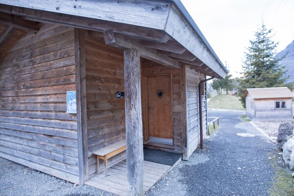 A wooden cabin with a wooden door, a bench outside, and a small building in the background. - Hüttenhotel Husky Lodge (Muotathal)