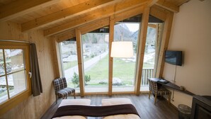 A bedroom with a large window, a bed, a chair, a desk, and a TV.