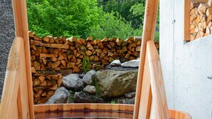 A wooden hot tub with a stone wall and logs in the background.