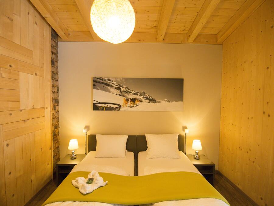 A hotel room with wooden walls, a bed with a green bedspread, two bedside tables with lamps, and a mountain landscape painting on the wall.