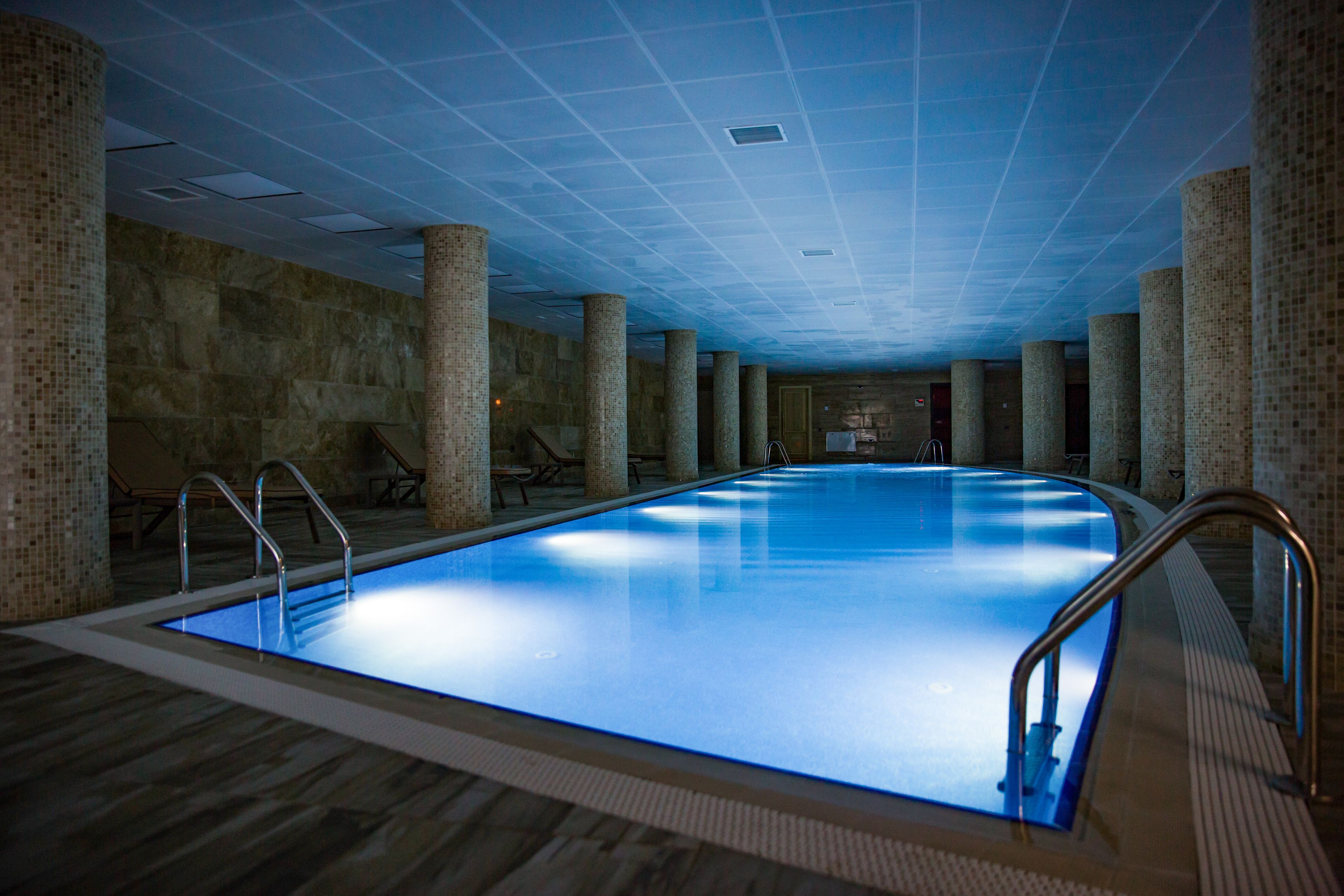 A large indoor swimming pool with tiled walls and columns, surrounded by seating areas.