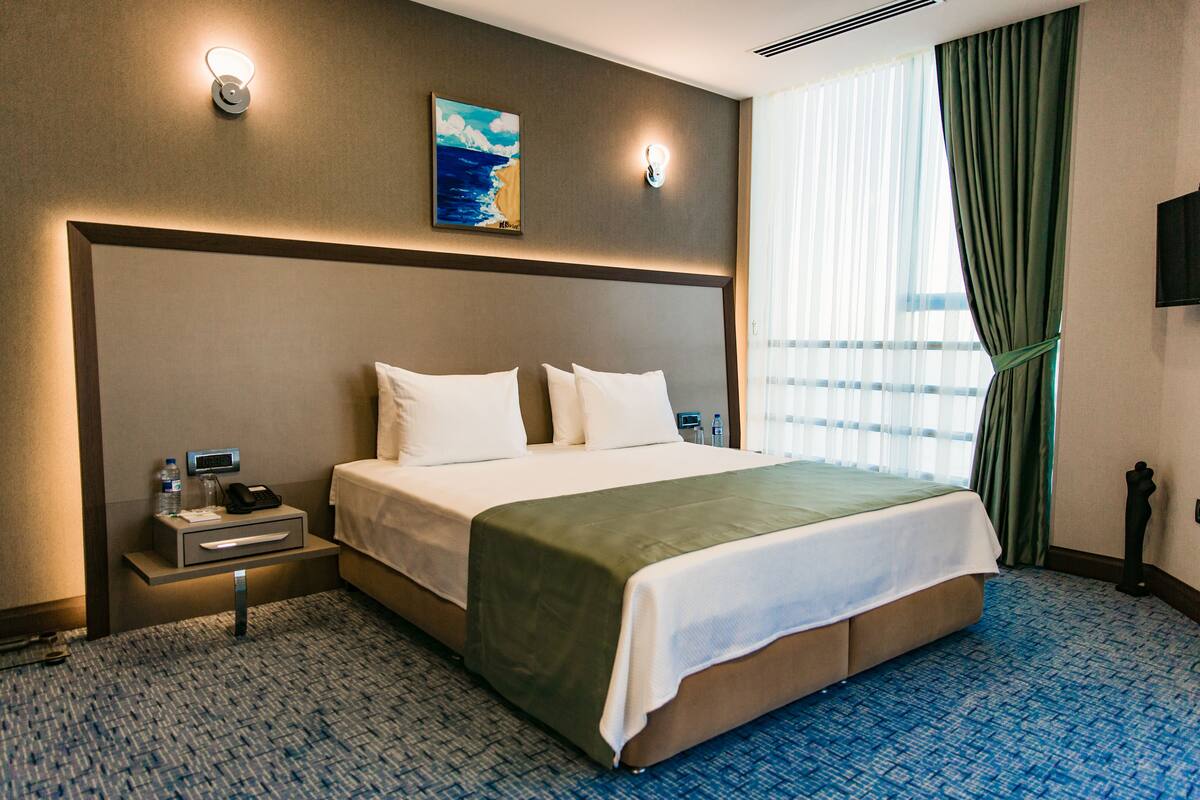 A hotel room with a large bed, a bedside table with a phone and water bottle, a painting on the wall, and a TV mounted on the wall.