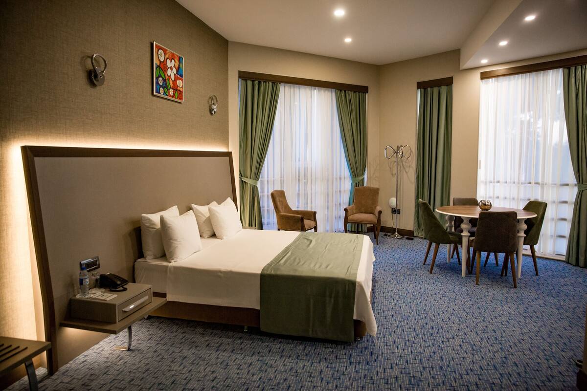 A hotel room with a large bed, a desk with a phone and a lamp, a chair, a small table with a globe, and two windows with green curtains.