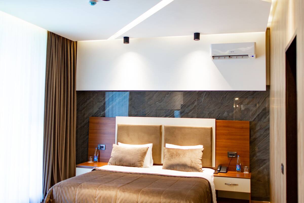 A modern hotel room with a large bed, wooden flooring, a marble accent wall, and a built-in air conditioning unit.