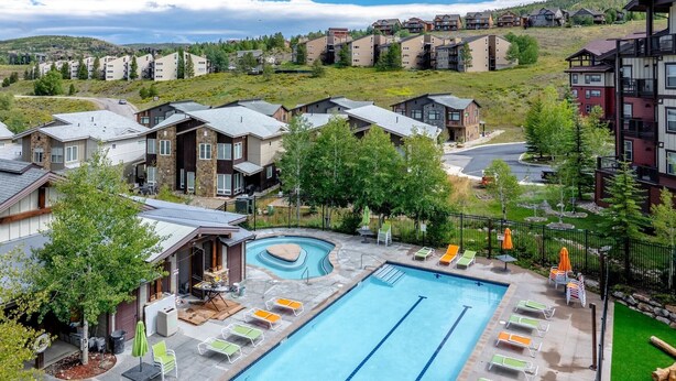 Piscina com várias pistas, rodeada por espreguiçadeiras e guarda-sóis, localizada numa área residencial com casas e um cenário montanhoso.