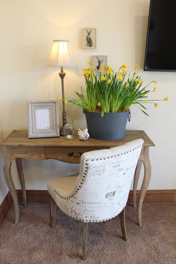 Un bureau en bois avec une lampe, un tableau encadré, un pot de fleurs avec des fleurs jaunes et une chaise au design vintage.