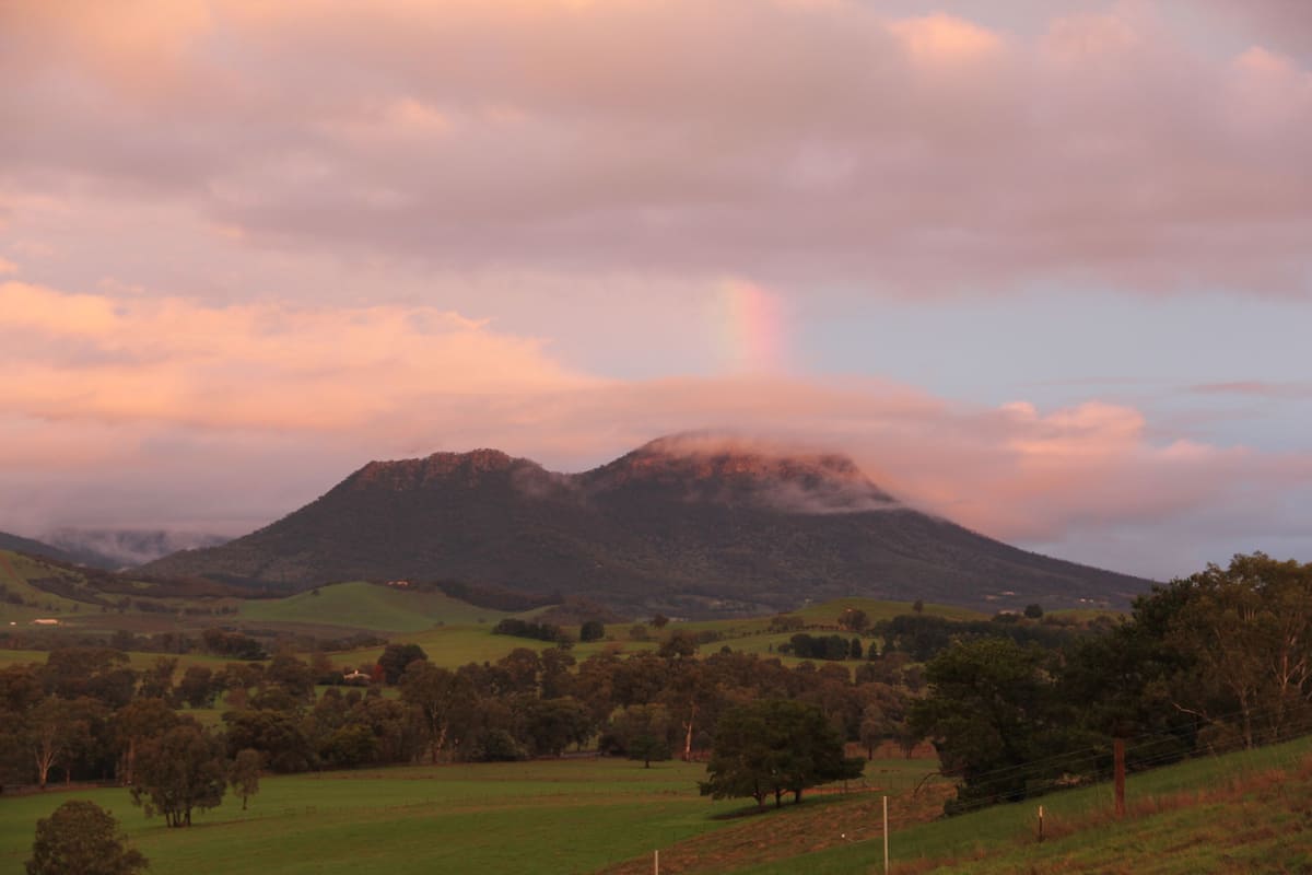 A mountain landscape with a rainbow, green fields, and scattered trees.