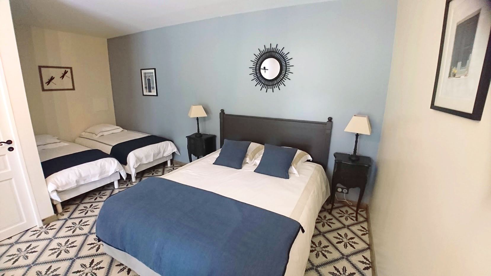 A hotel room with a large bed, two smaller beds, a wall clock, a sunburst mirror, and patterned floor tiles.