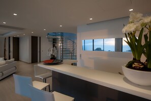 A modern interior with a white reception desk, a white sofa, and a dining area. There is a large window with a view of the sea.