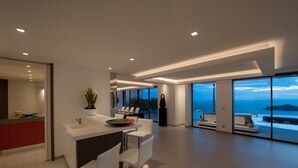 Une cuisine moderne avec un plan de travail blanc, un évier et un coin repas avec des chaises blanches. Une grande fenêtre offre une vue sur l’océan, et un balcon aménagé avec des sièges est également présent.