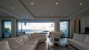 Un salon moderne avec un grand canapé d’angle blanc, une table basse et une vue sur l’océan grâce à de grandes fenêtres.
