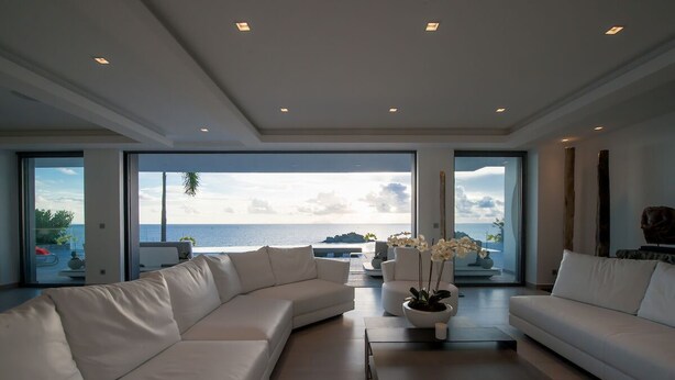 Un salon moderne avec un grand canapé d’angle blanc, une table basse et une vue sur l’océan grâce à de grandes fenêtres.