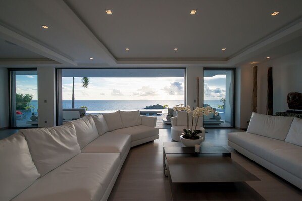A modern living room with a large white sectional sofa, a coffee table, and a view of the ocean through large windows.