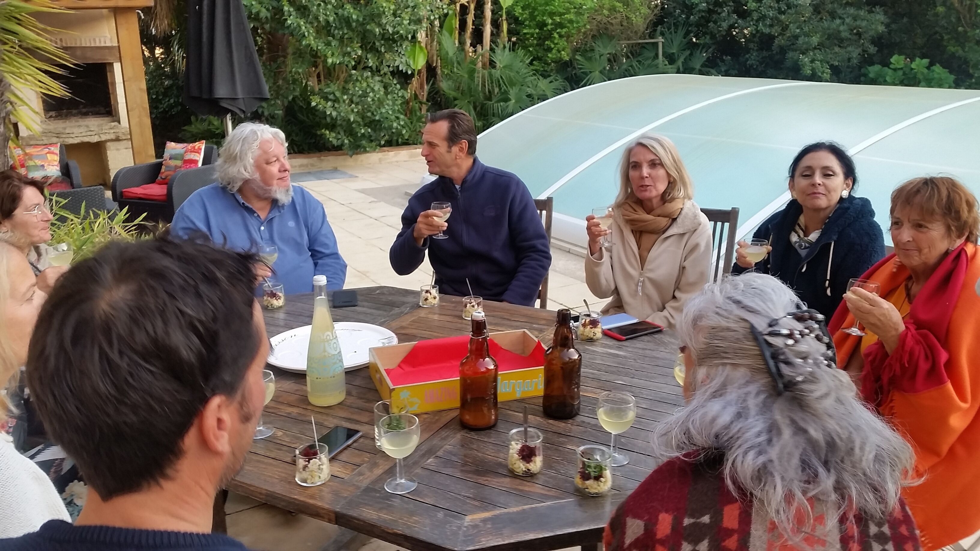 Un grupo de personas sentadas alrededor de una mesa de madera al aire libre, con bebidas y una botella de agua.