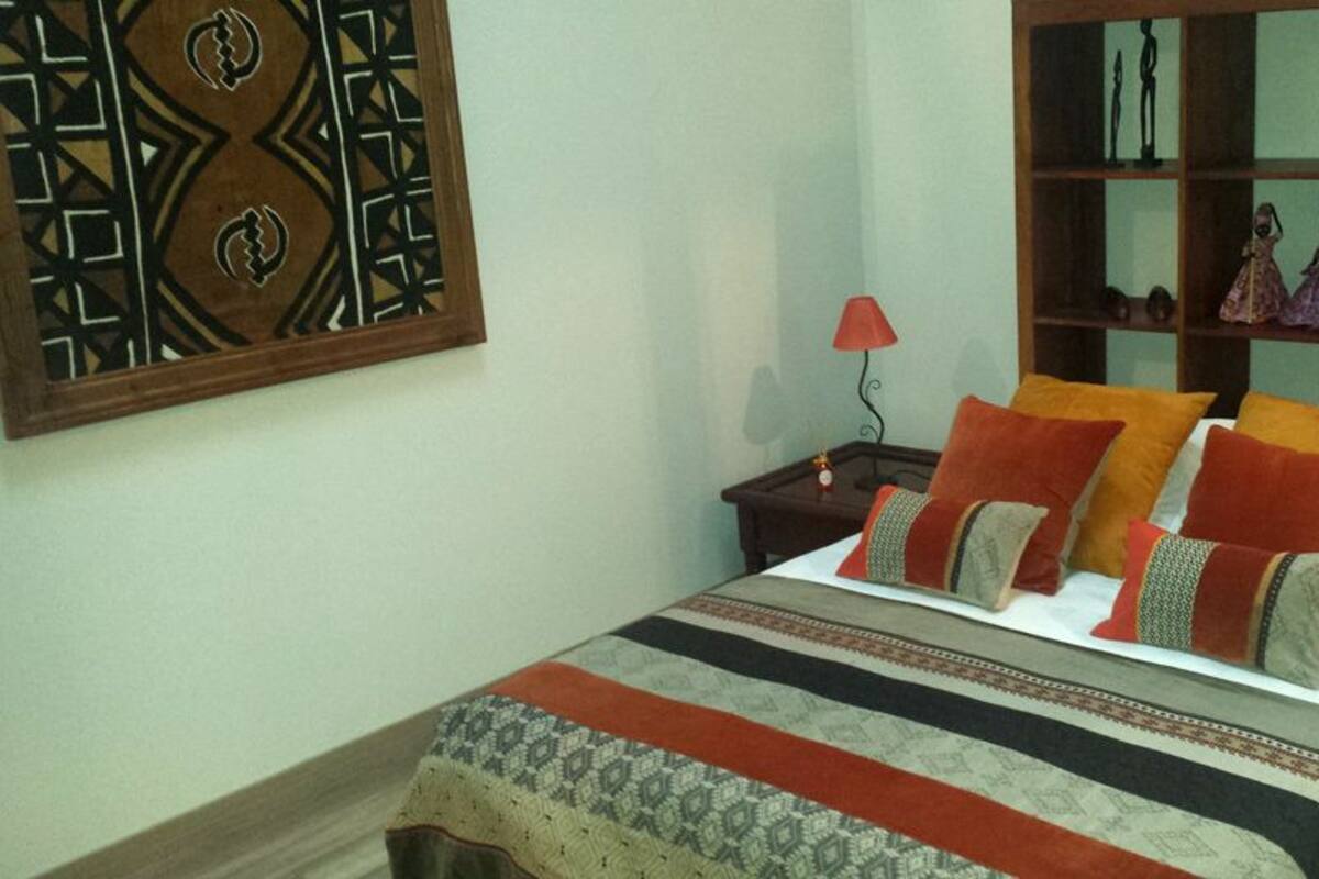 A neatly made bed with a patterned bedspread and multiple pillows, a nightstand with a lamp, and a shelf with decorative items.